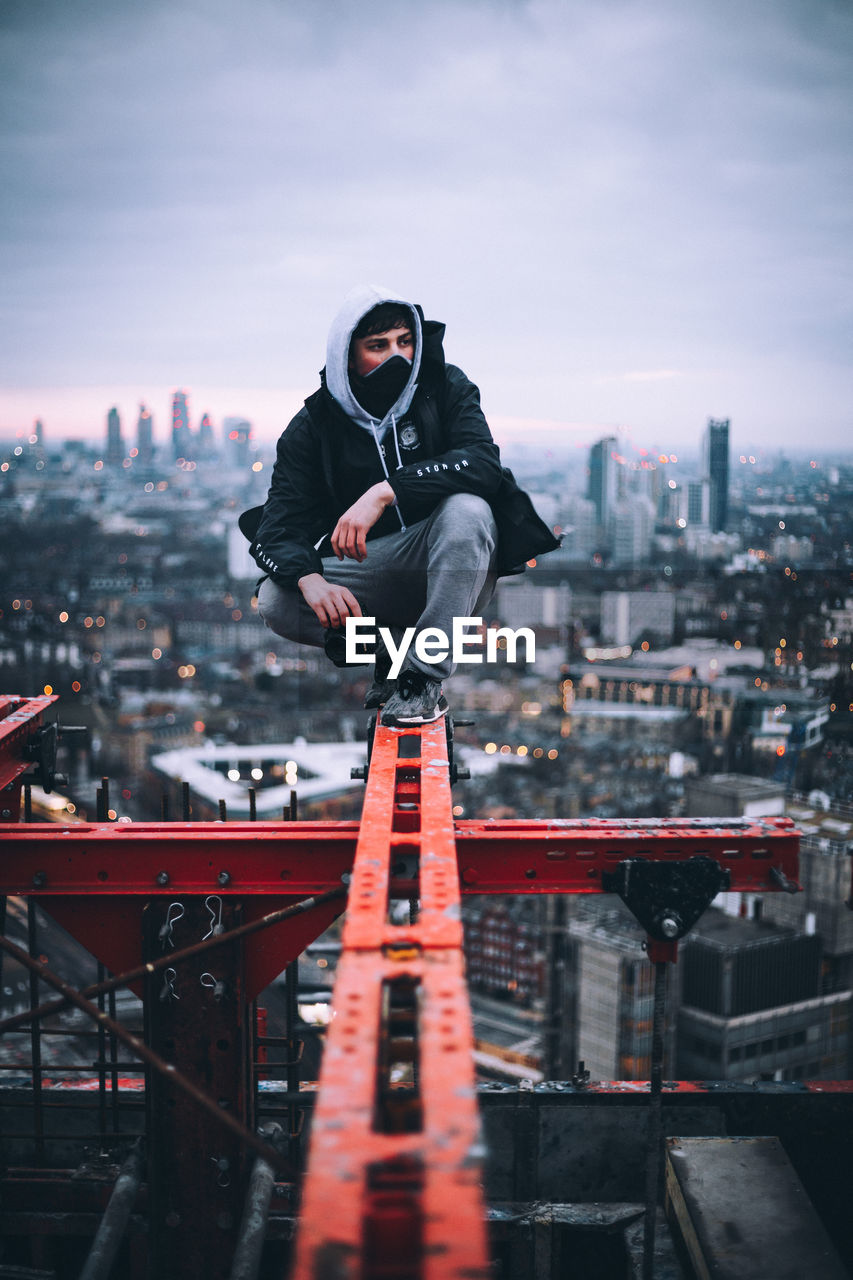 Young man looking away while sitting on metal railing against sky