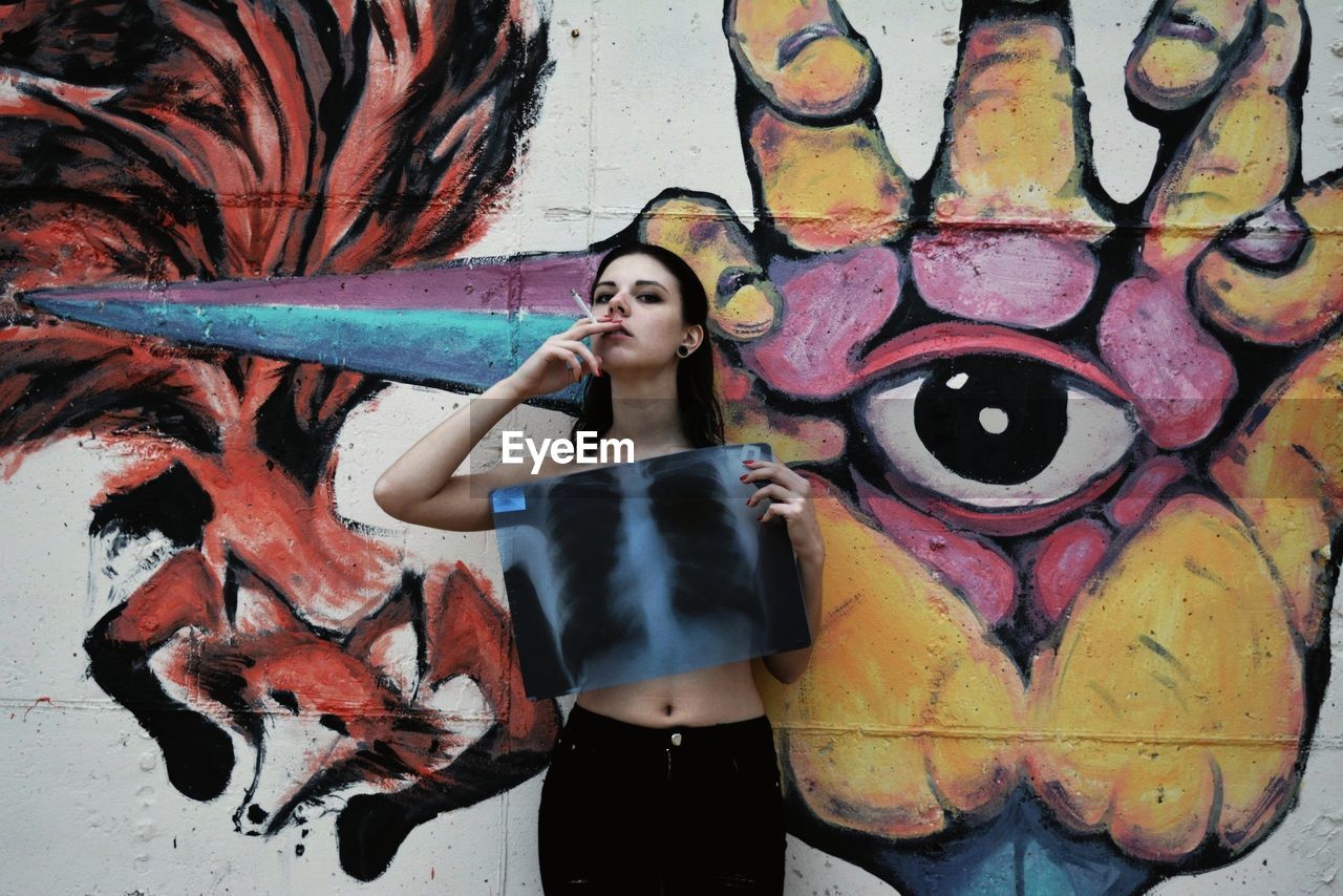 Young woman smoking cigarette and covering breast with medical x-ray while standing against graffiti wall