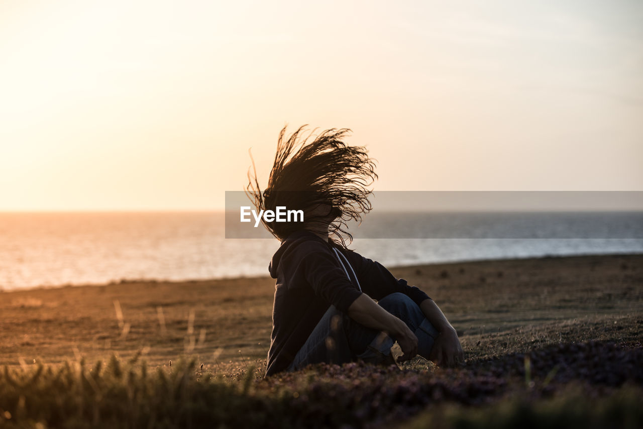 WOMAN SITTING AT BEACH AGAINST SKY DURING SUNSET