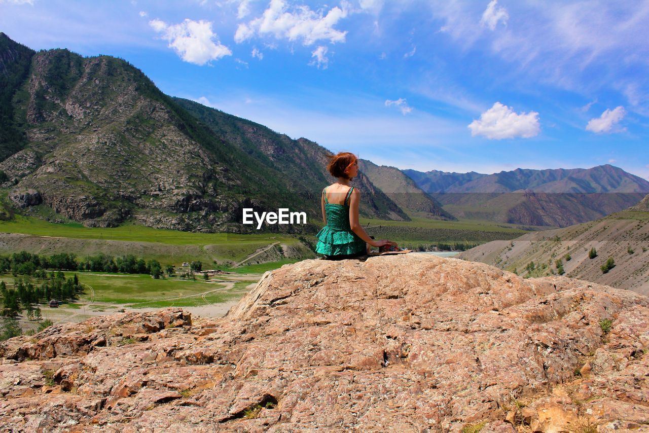 Rear view of young woman sitting on rock against mountains