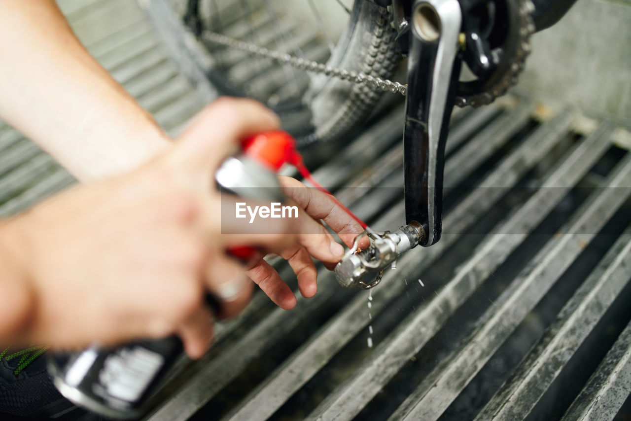 From above of crop anonymous male master using chemical spray and cleaning bike pedal in repair workshop