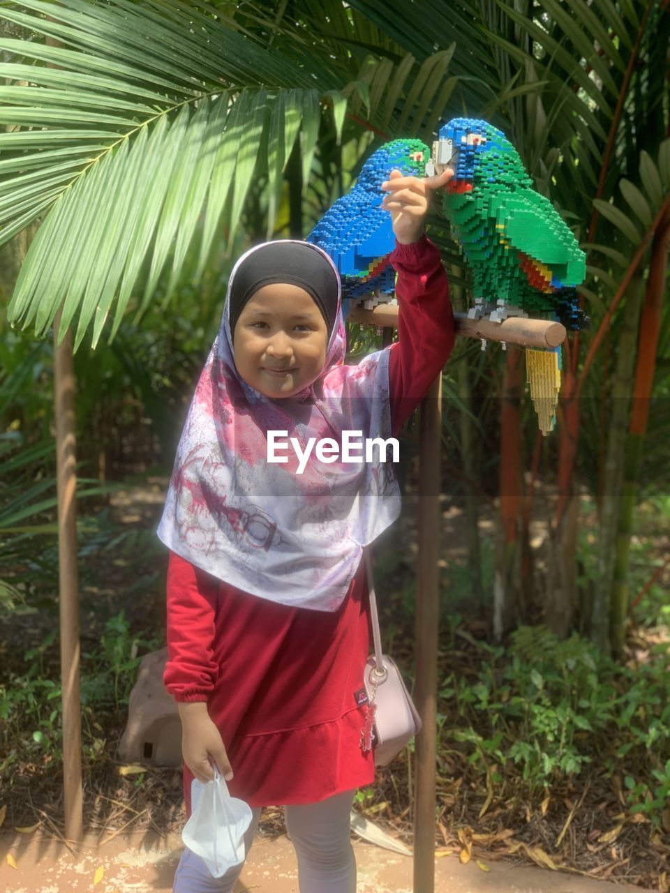 child, childhood, full length, female, women, nature, standing, smiling, tree, plant, clothing, day, men, front view, tropical climate, leisure activity, lifestyles, happiness, one person, looking at camera, portrait, person, adult, outdoors, palm tree, jungle, human face, emotion, holding, casual clothing, traditional clothing, agriculture