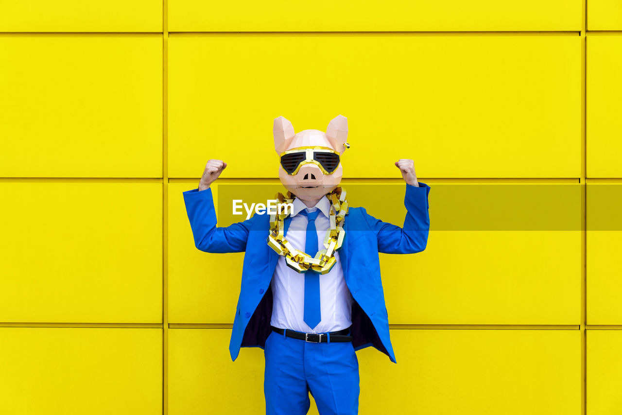 Man wearing vibrant blue suit, pig mask and large golden chain flexing muscles against yellow wall
