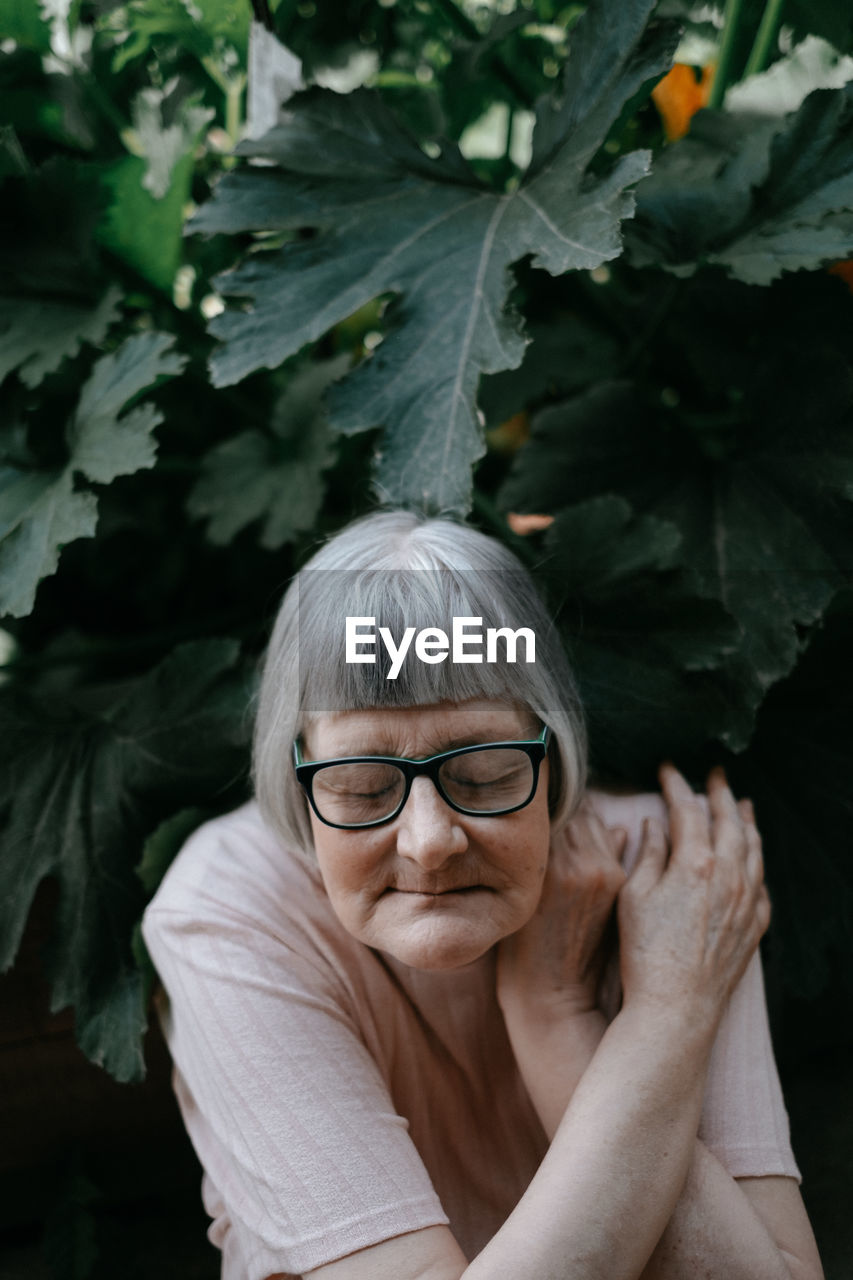 Senior woman with grey hair and closed eyes in eyeglasses crossing arms over shoulder among plants with big green leafs