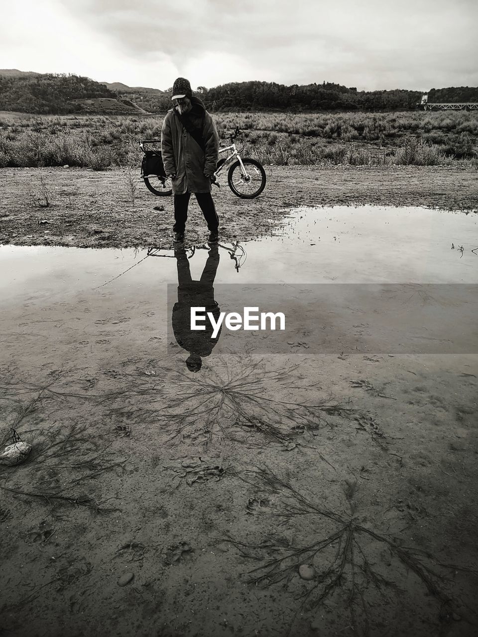 black and white, water, monochrome, bicycle, one person, monochrome photography, sky, black, nature, sea, land, men, adult, full length, white, cloud, reflection, environment, beach, activity, lifestyles, sports, leisure activity, landscape, day, person, outdoors, sand, darkness, coast, transportation, wave, wet, vehicle, puddle, scenics - nature