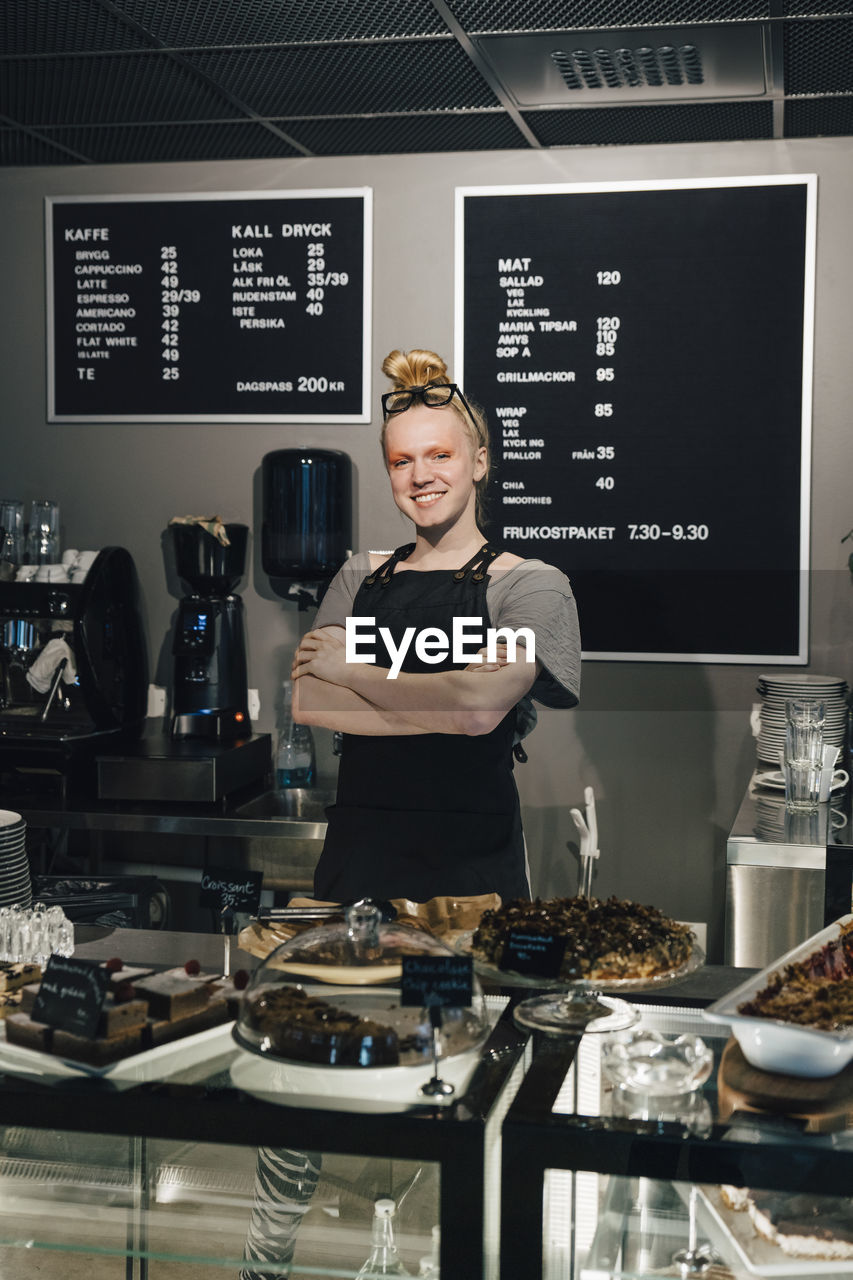 Portrait of smiling barista standing with arms crossed by food variation on counter in illuminated cafeteria