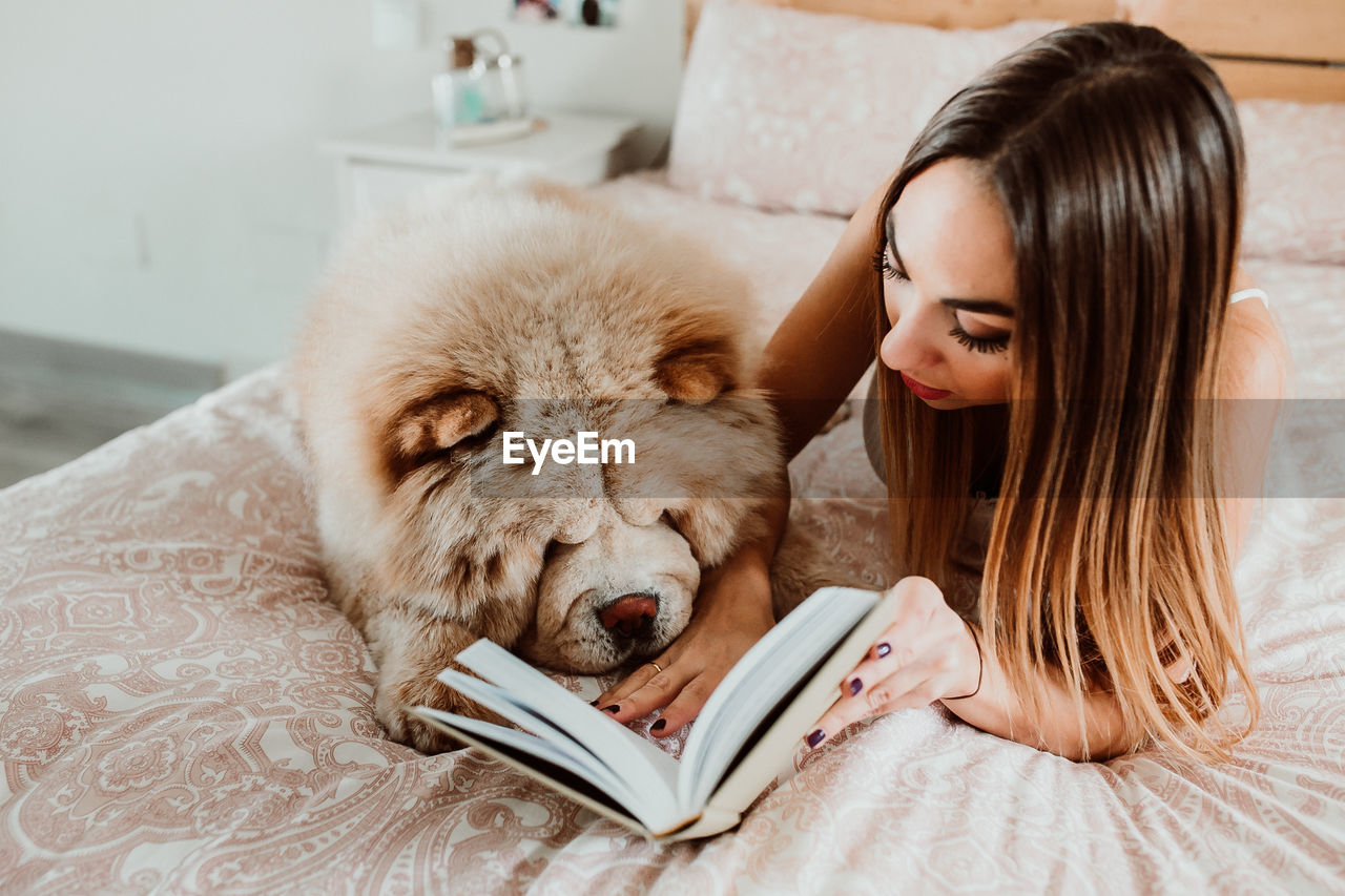 Young woman reading book while lying with dog on bed