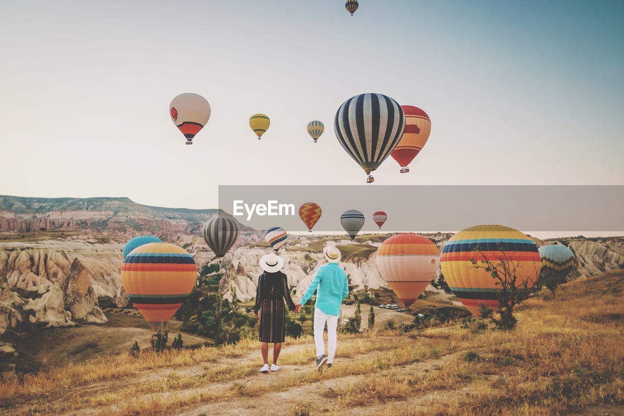 Rear view of couple looking at hot air balloons flying over land against sky