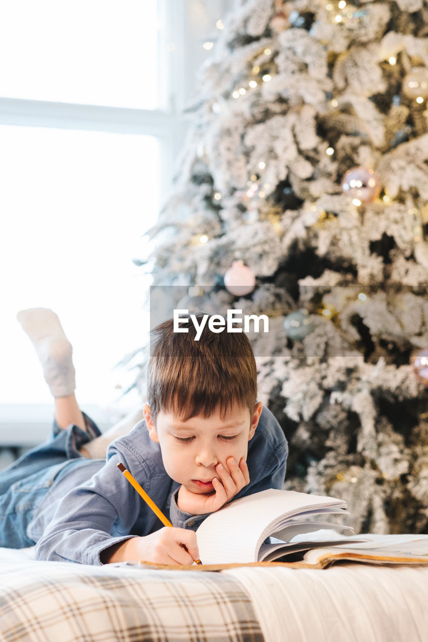 Boy doing homework lying on the sofa, against the backdrop of a christmas tree, in his room