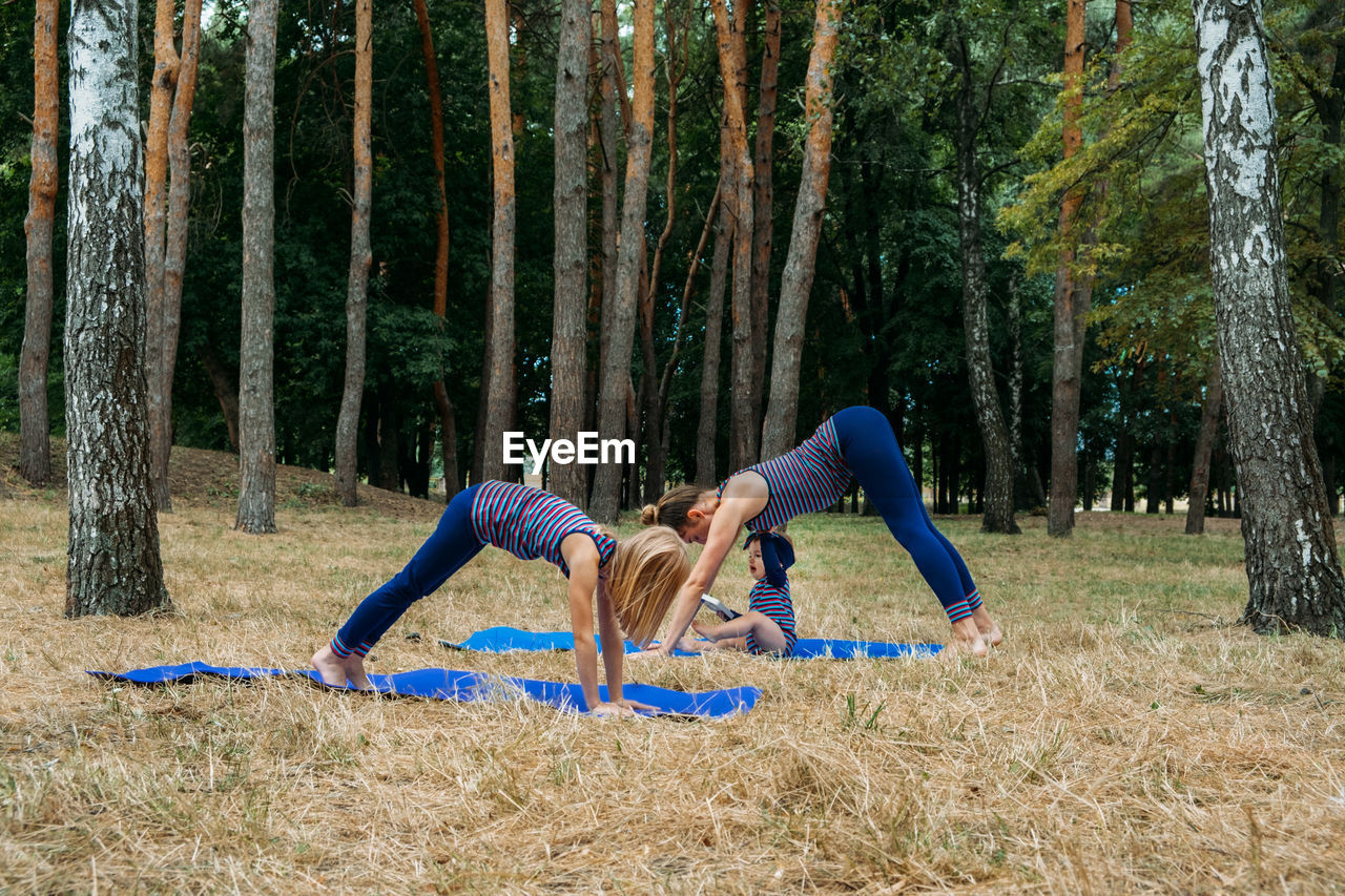 Family fitness and yoga. happy mom exercising with baby girls on grassy land against trees in