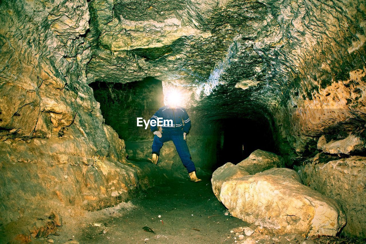 REAR VIEW OF MAN STANDING ON ROCK BY CAVE