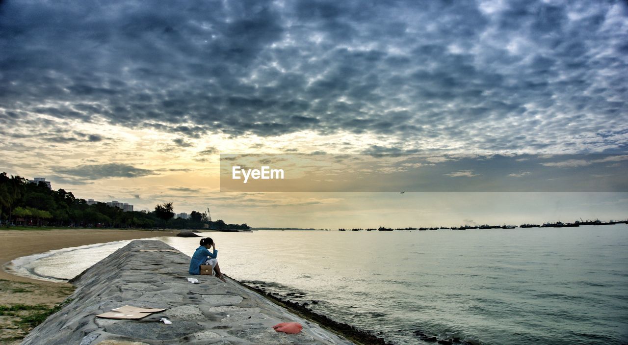 Woman sitting on stone pier in front of sea against cloudy sky