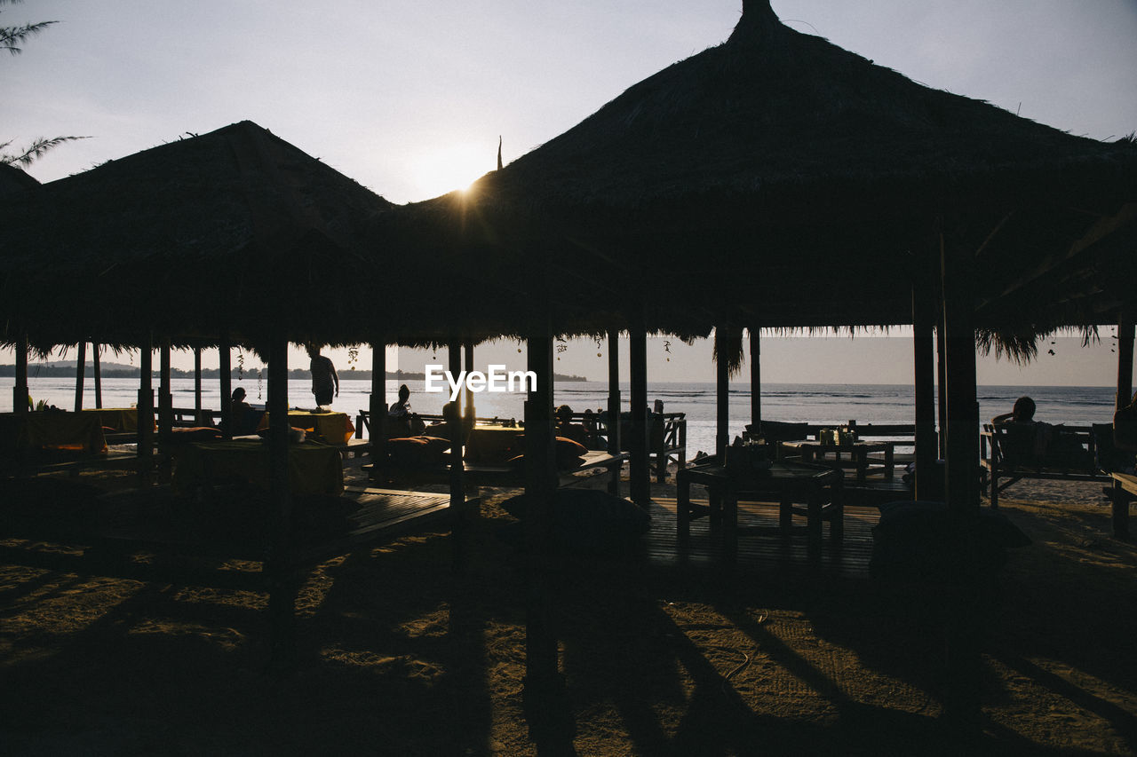 Silhouette thatched roof huts at beach against sky