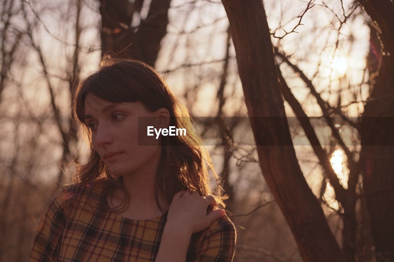 Woman looking away against bare trees during sunset