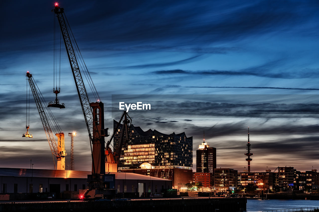 Cranes and buildings in city against cloudy sky at sunset