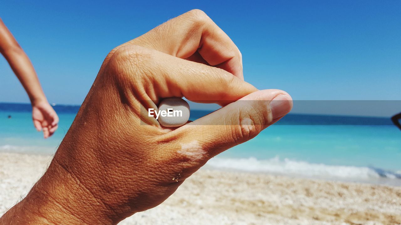 Close-up of man hand holding sand at beach against clear blue sky