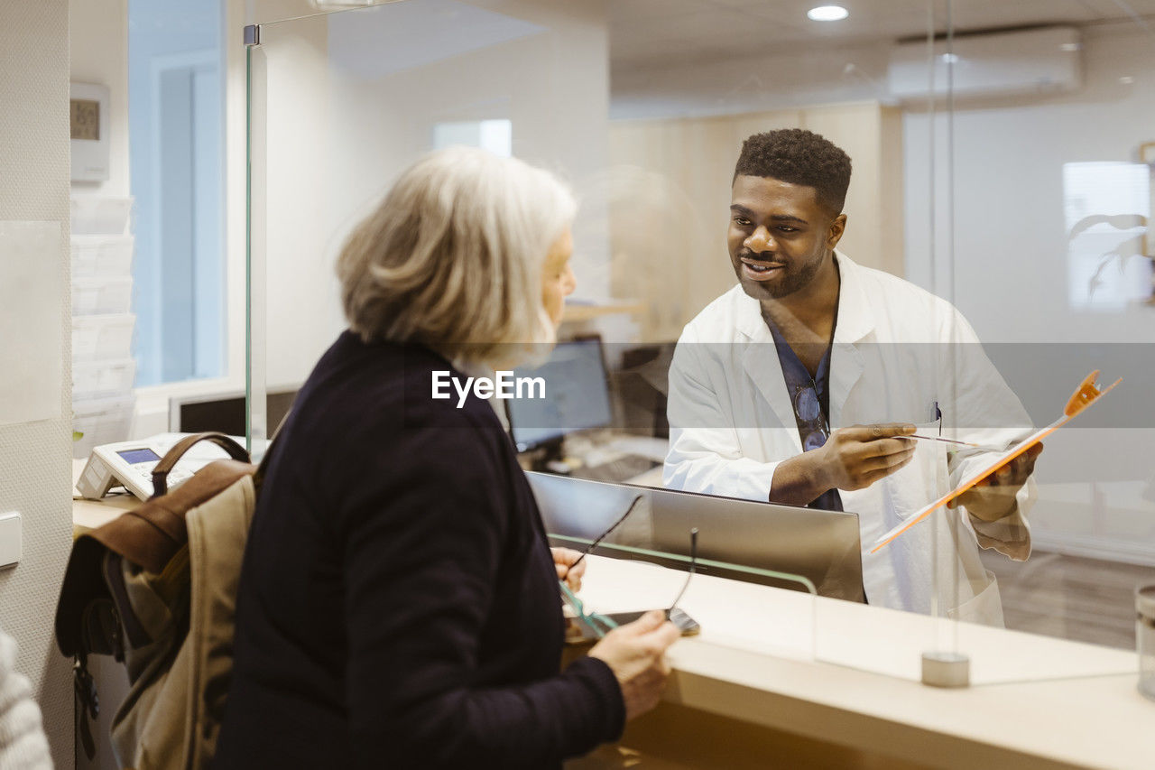 Smiling male receptionist holding clipboard while talking to senior woman through transparent shield in clinic