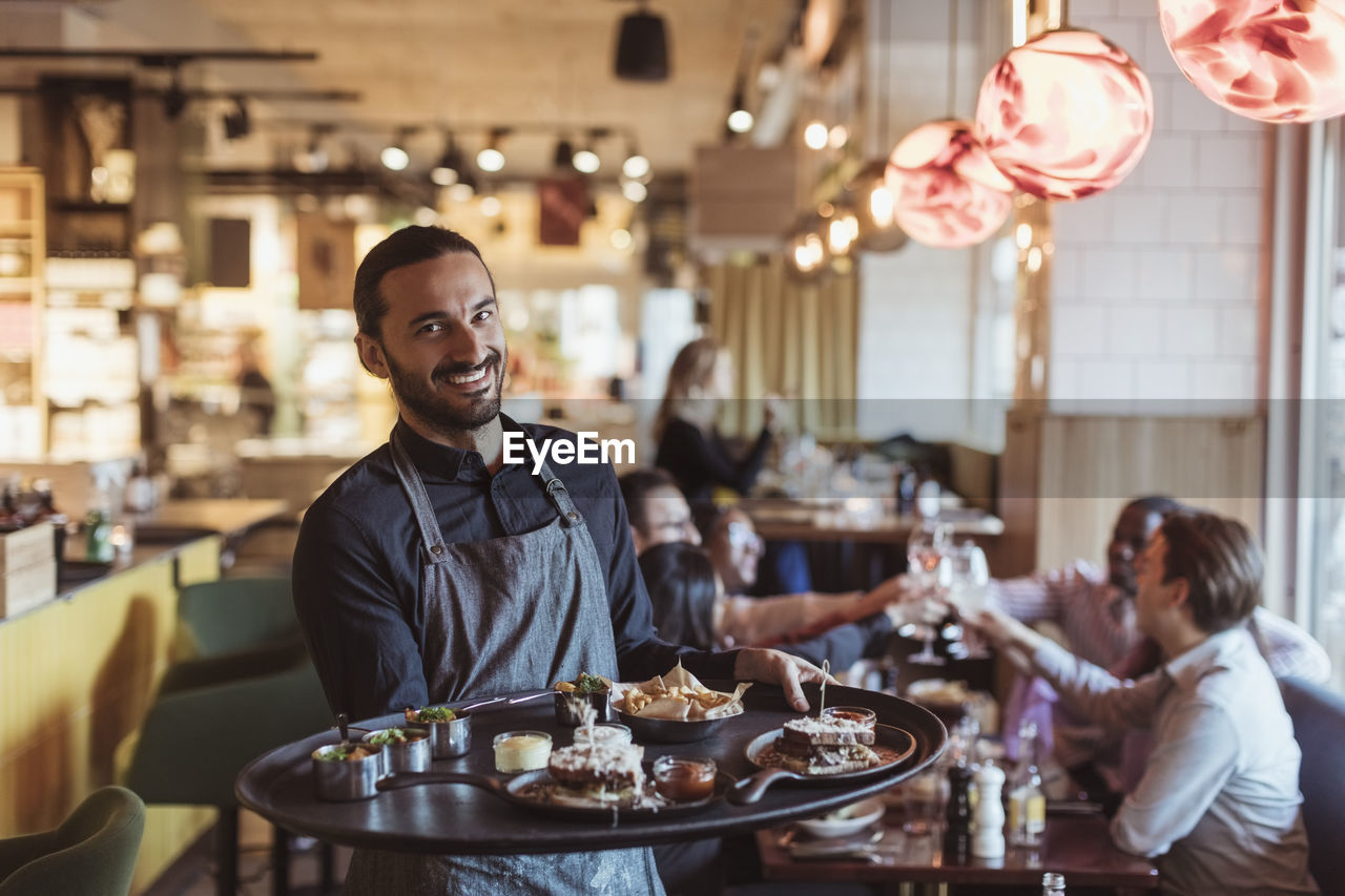 Portrait of male waiter with food tray while customers toasting in background at bar