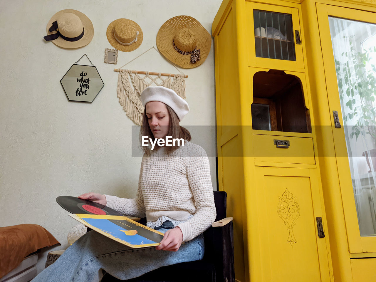 Young woman in beret sits in cozy home, room with music record in hands. vintage yellow cabinet.