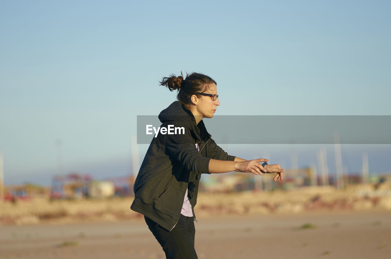 Side view of young woman gesturing while standing at beach against clear blue sky