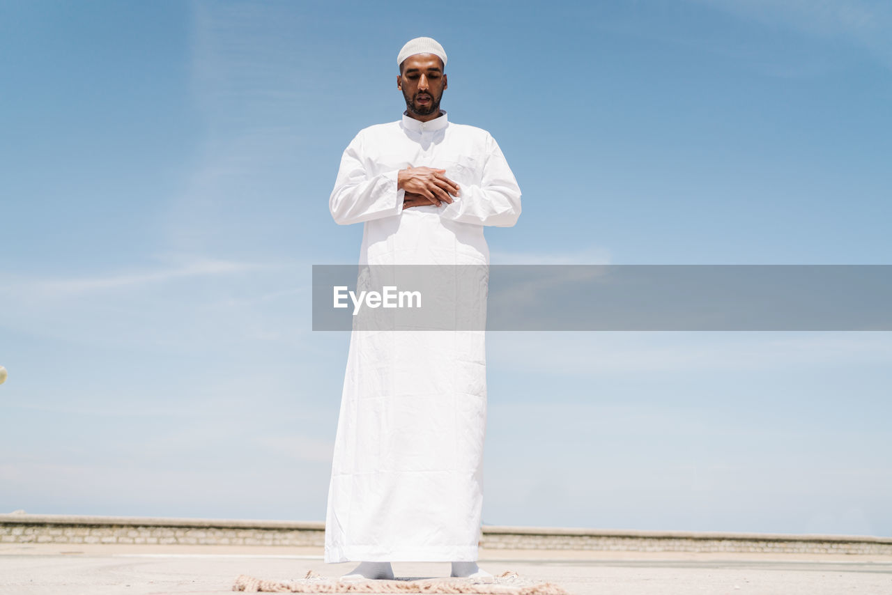 Full boy islamic male in traditional white clothes standing on rug and praying against blue sky on beach