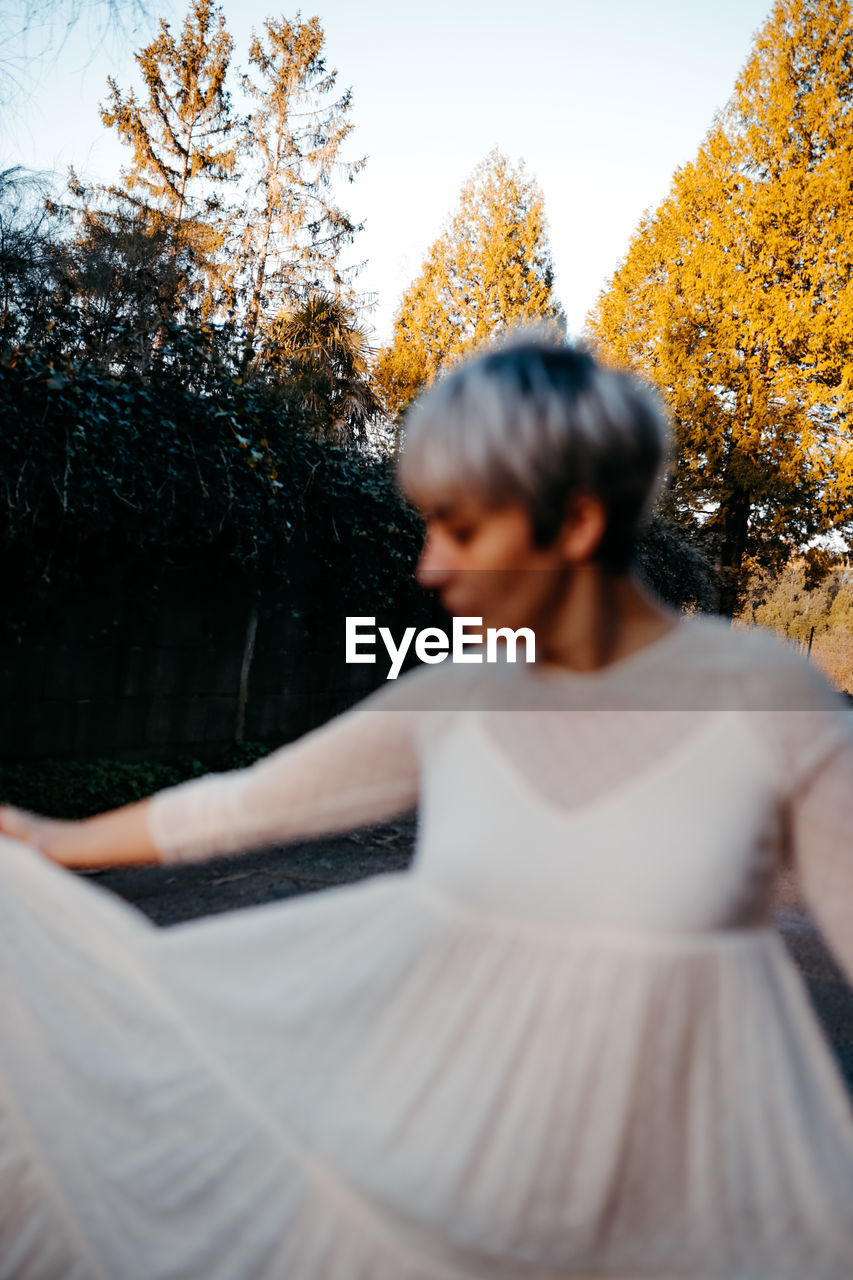 Side view of blurred anonymous female with short hair wearing white dress standing against tall green trees in rural terrain