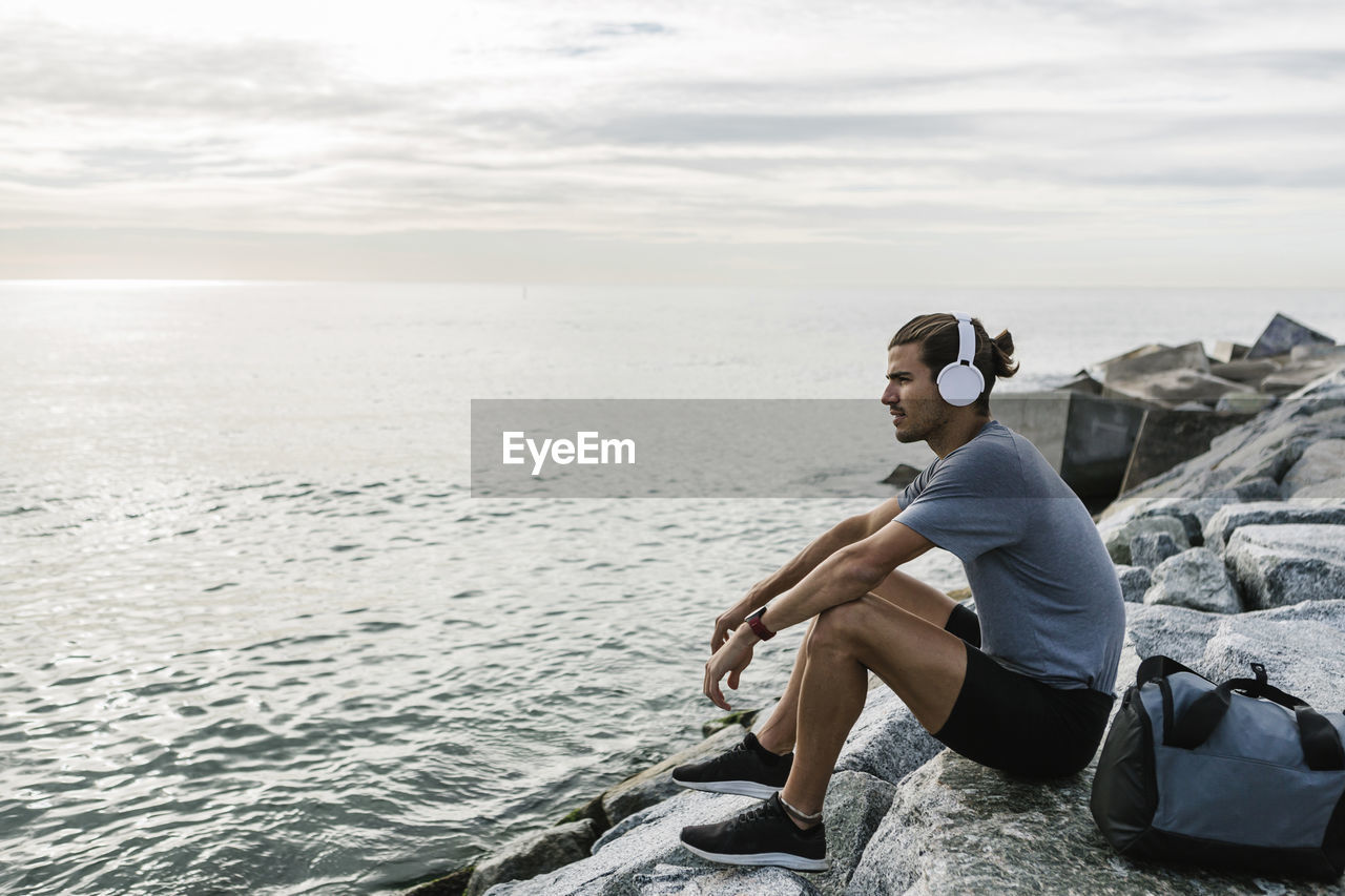 Sportsman with headphones looking at view while sitting on rock by sea