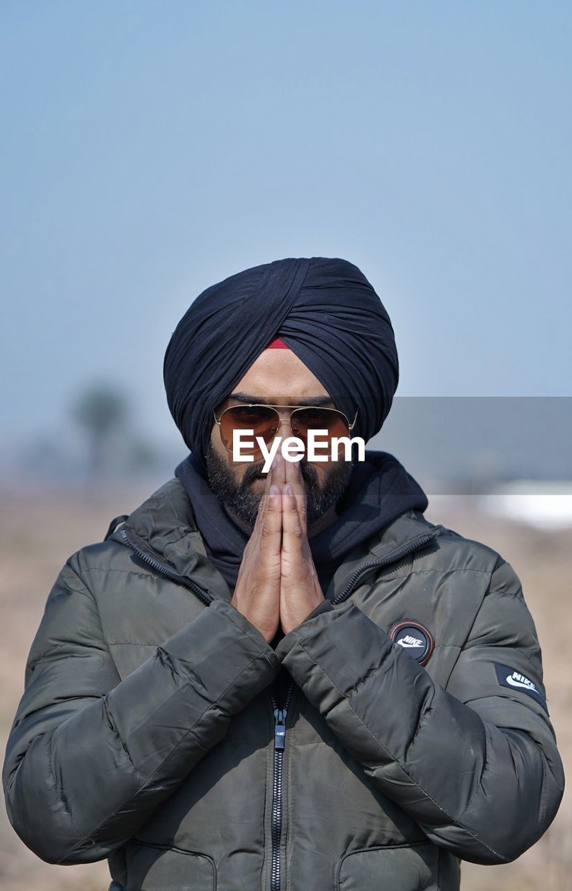 Young man wearing turban with hands clasped standing against clear sky