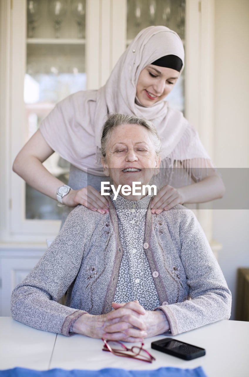 Young female home caregiver giving shoulder massage to grandmother at home