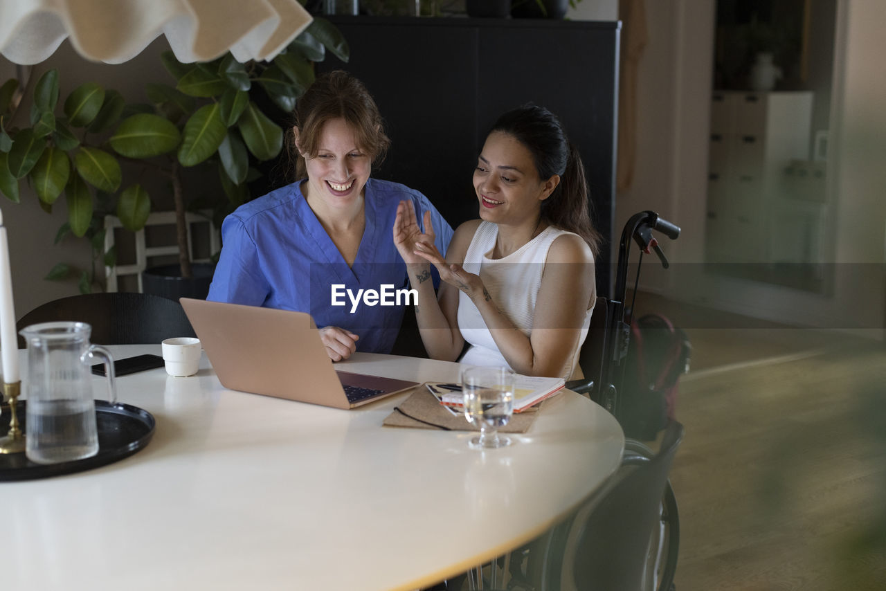 Woman with paraplegia gesturing by caregiver while watching laptop at home