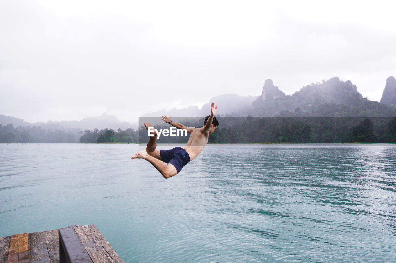 Shirtless man jumping in lake against sky