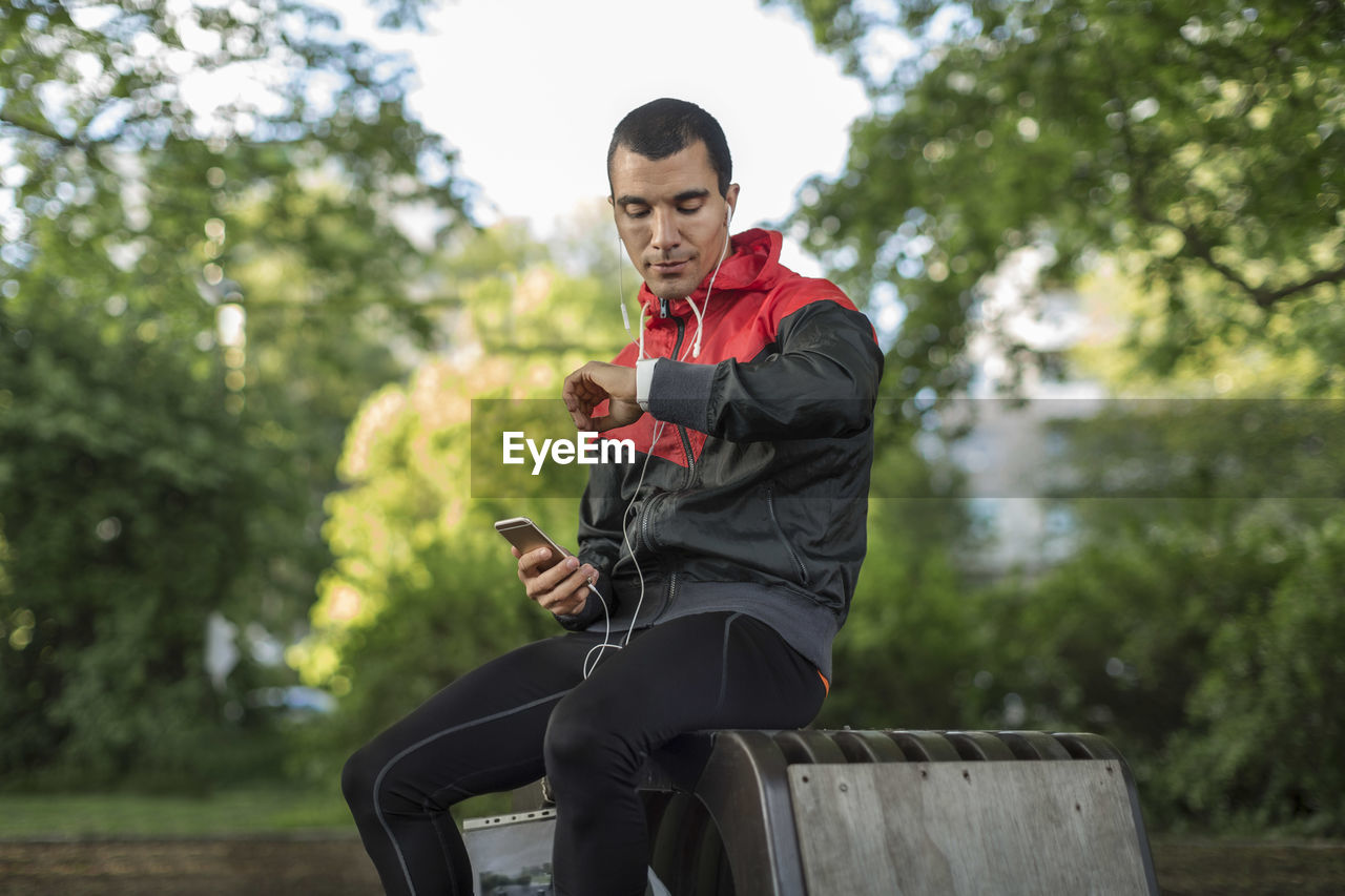 Man sitting on bench holding phone looking at wristwatch