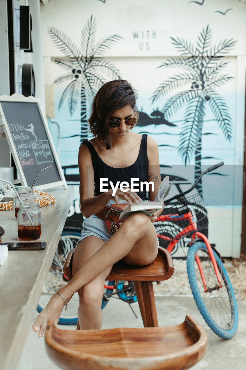 Resting woman in casual wear and trendy sunglasses reading book during refreshment in outdoors bar