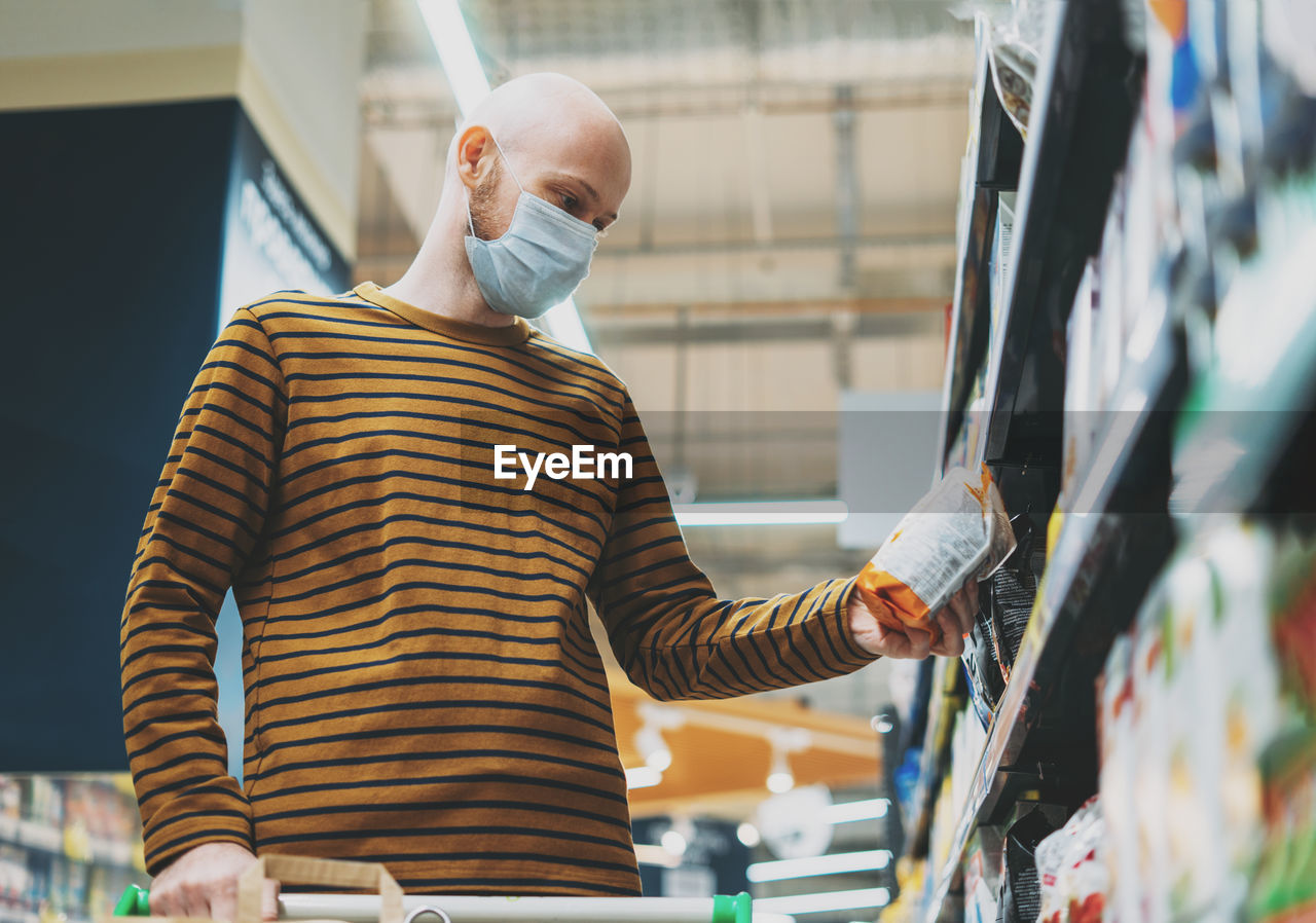 Bald man in medical face mask chooses products in supermarket, concept of coronavirus quarantine