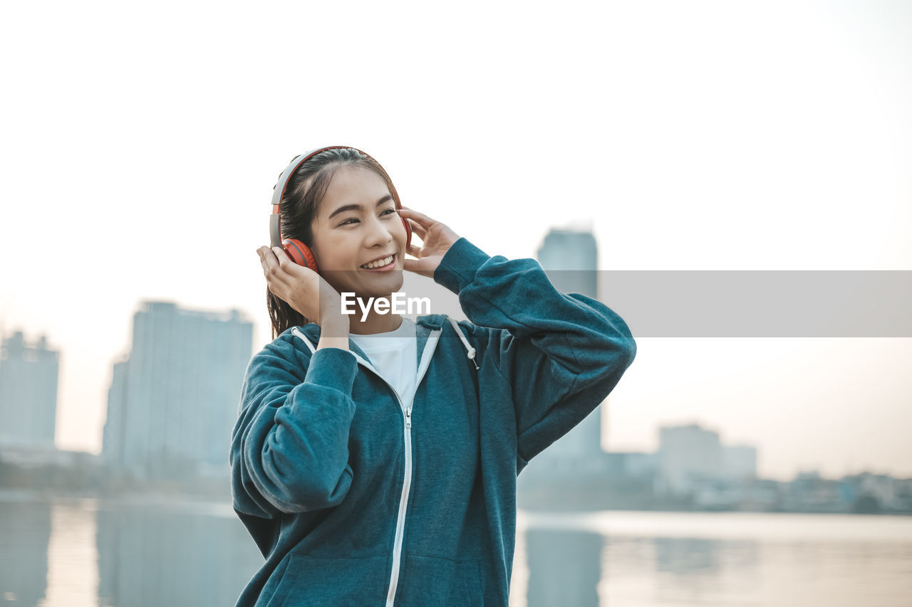 PORTRAIT OF SMILING YOUNG WOMAN STANDING AGAINST SKY