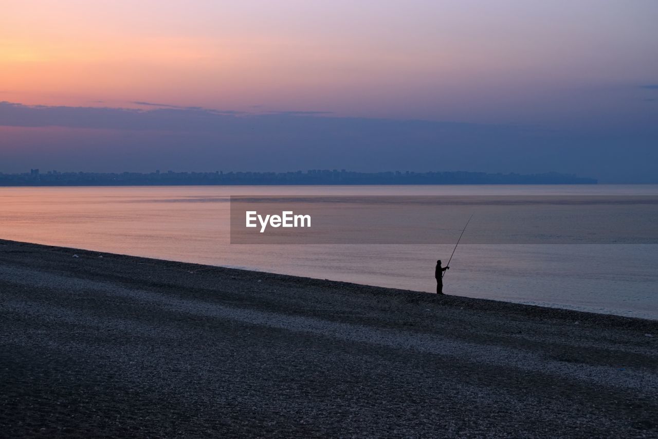 Man fishing on shore at beach during sunset