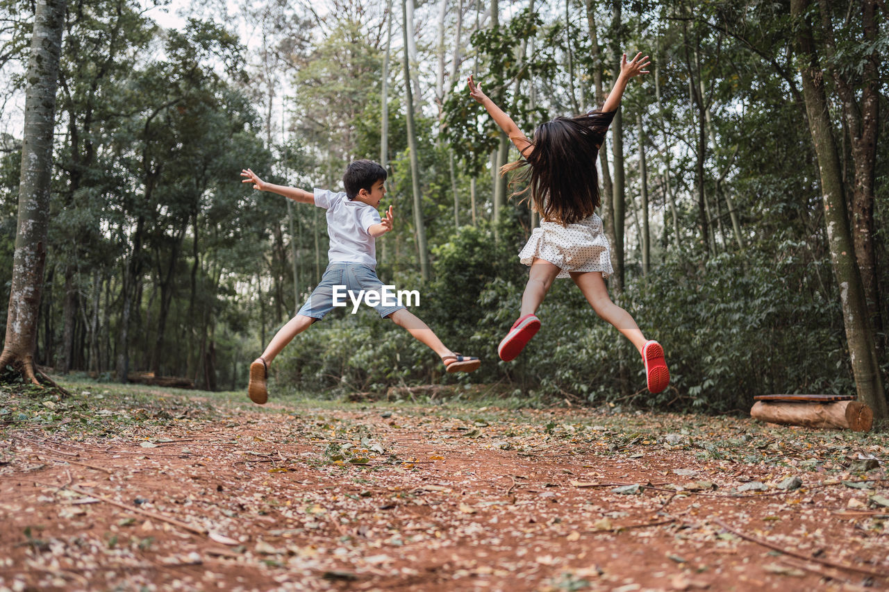 Back view of anonymous joyful boy with sister jumping with outstretched arms over terrain with trees