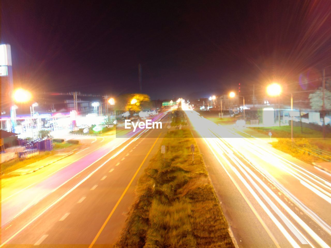 LIGHT TRAILS ON ROAD ALONG ILLUMINATED STREET AT NIGHT