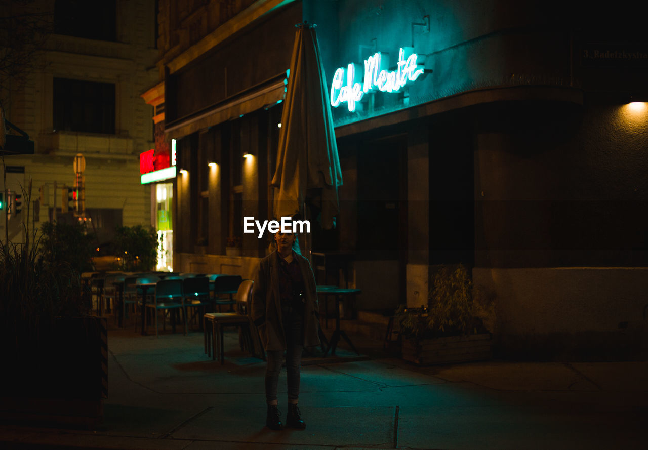 REAR VIEW OF MAN WALKING ON ILLUMINATED STREET AGAINST BUILDINGS AT NIGHT
