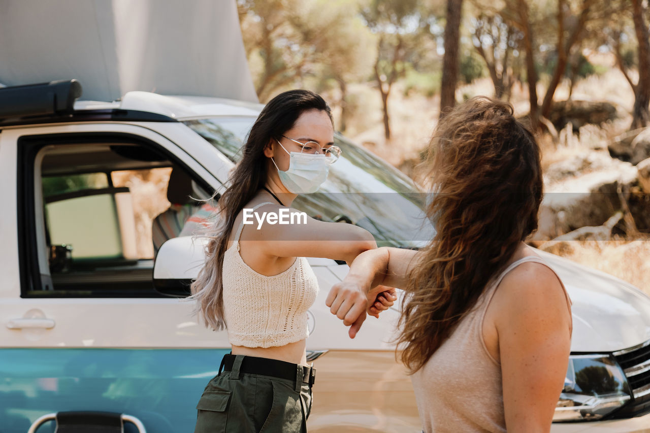 Friendly women in protective masks standing near van in forest and bumping elbows while greeting each other during coronavirus epidemic