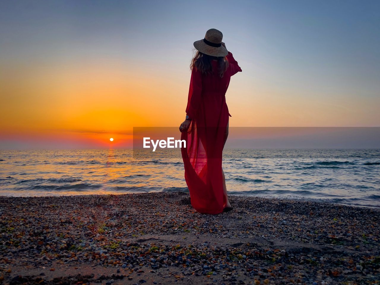 Rear view of woman wearing long red dress standing alone on the beach at sunrise