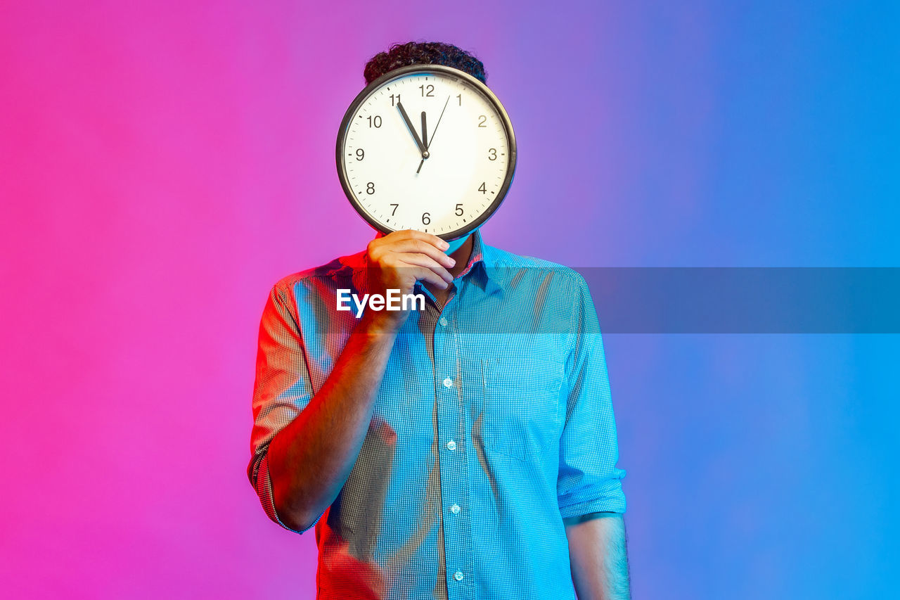 Man holding clock in front of face against colored background