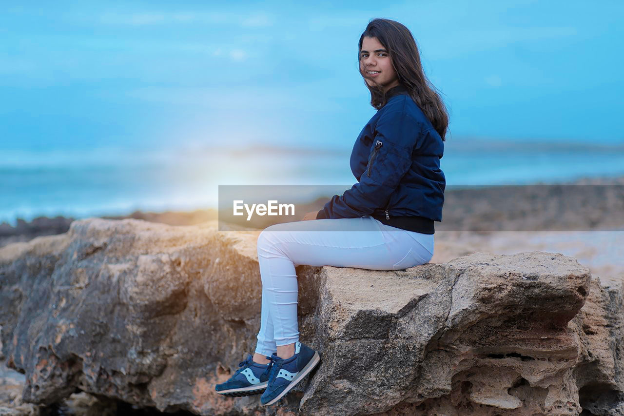 YOUNG MAN SITTING ON ROCK AGAINST SEA