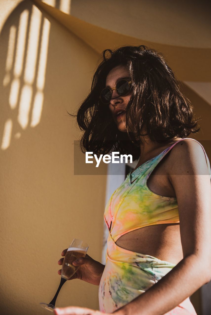 Young woman wearing sunglasses standing against wall at home