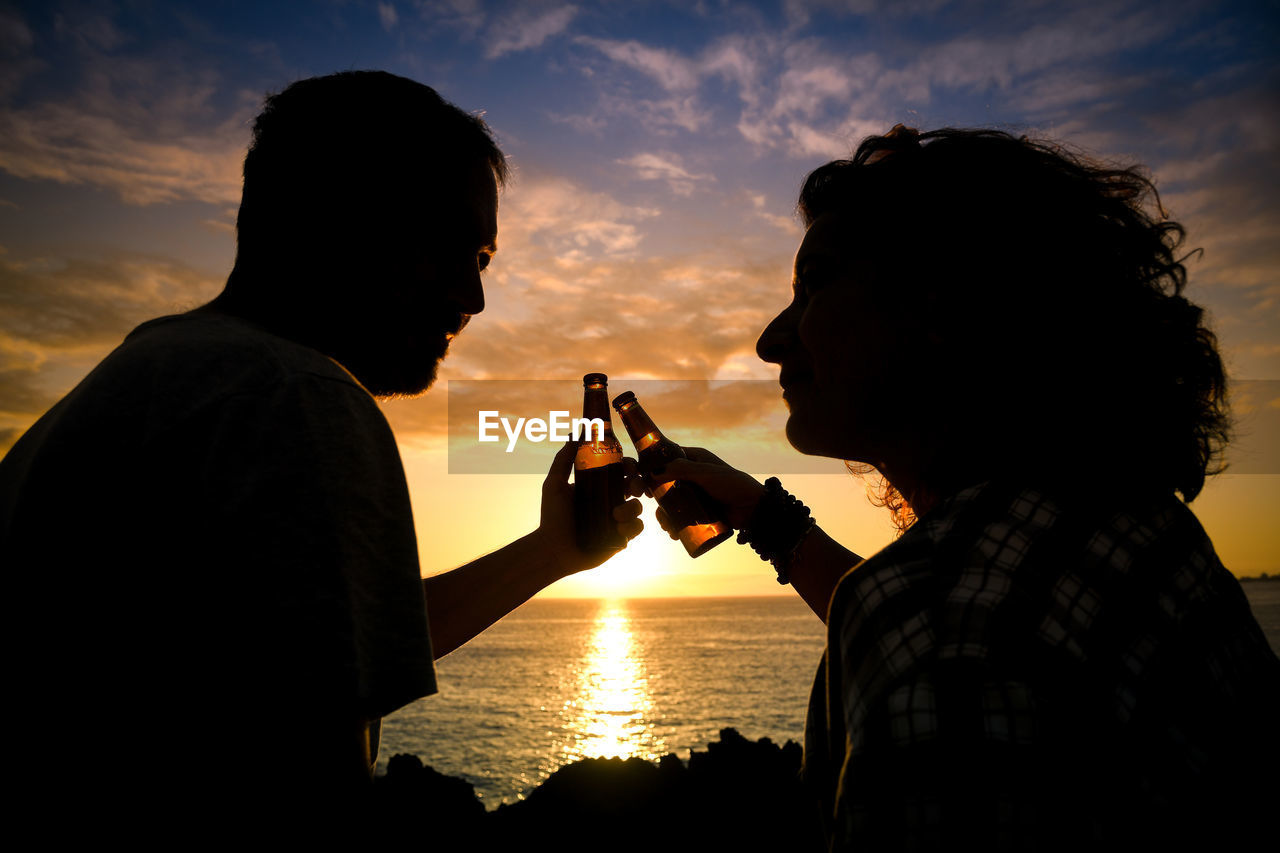Couple toasting beer bottles while sitting by sea against sky during sunset