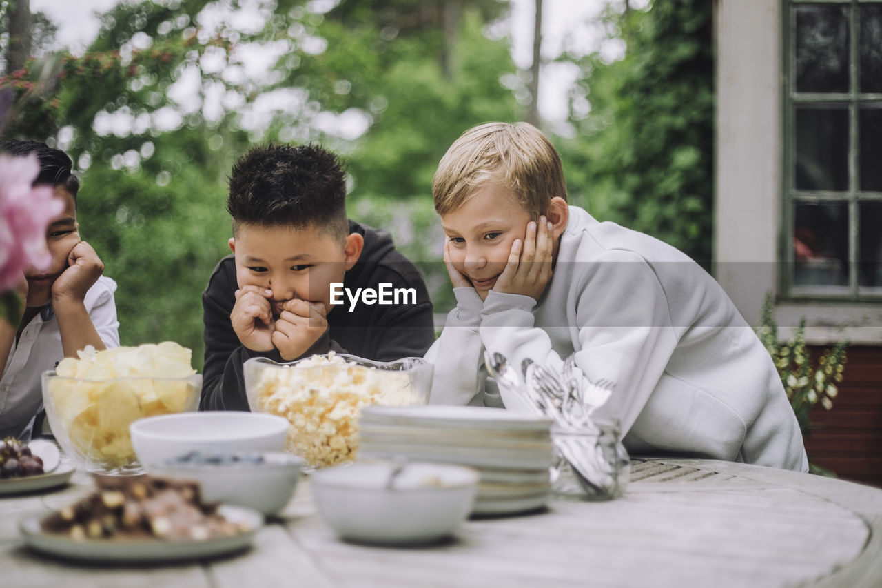 Curious boys leaning on elbows while looking at snacks bowl on table