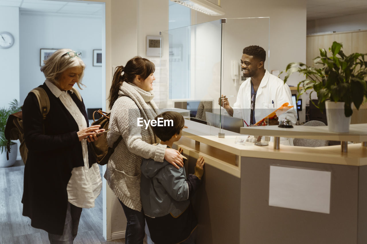 Patients standing and talking to male receptionist through transparent shield in clinic