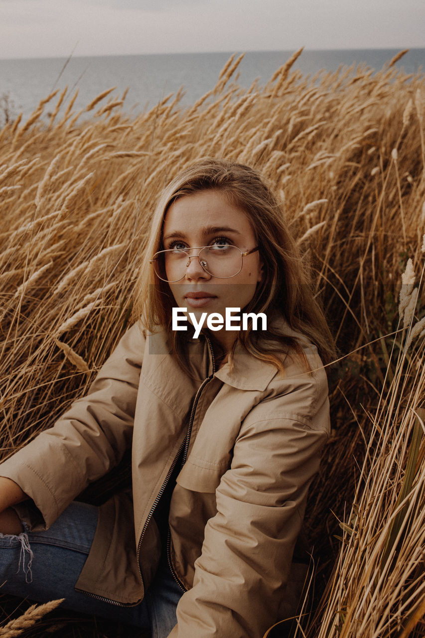 Young woman looking away while sitting by plants against sky