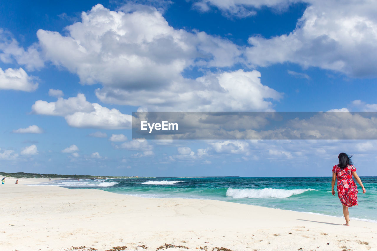 Rear view of mid adult woman walking at beach against cloudy sky