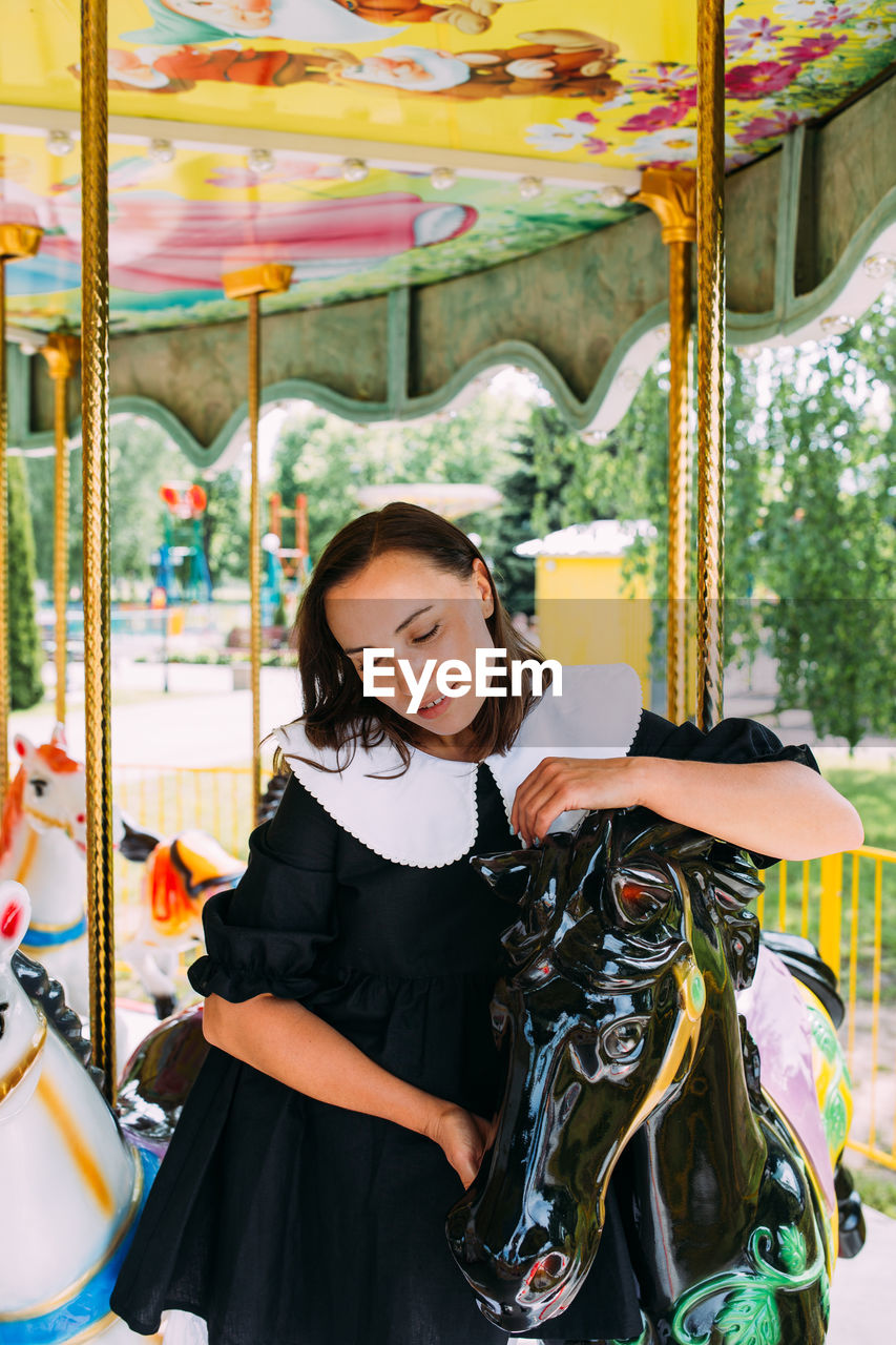 Beautiful brunette girl in a black dress poses on a bright carousel with horses